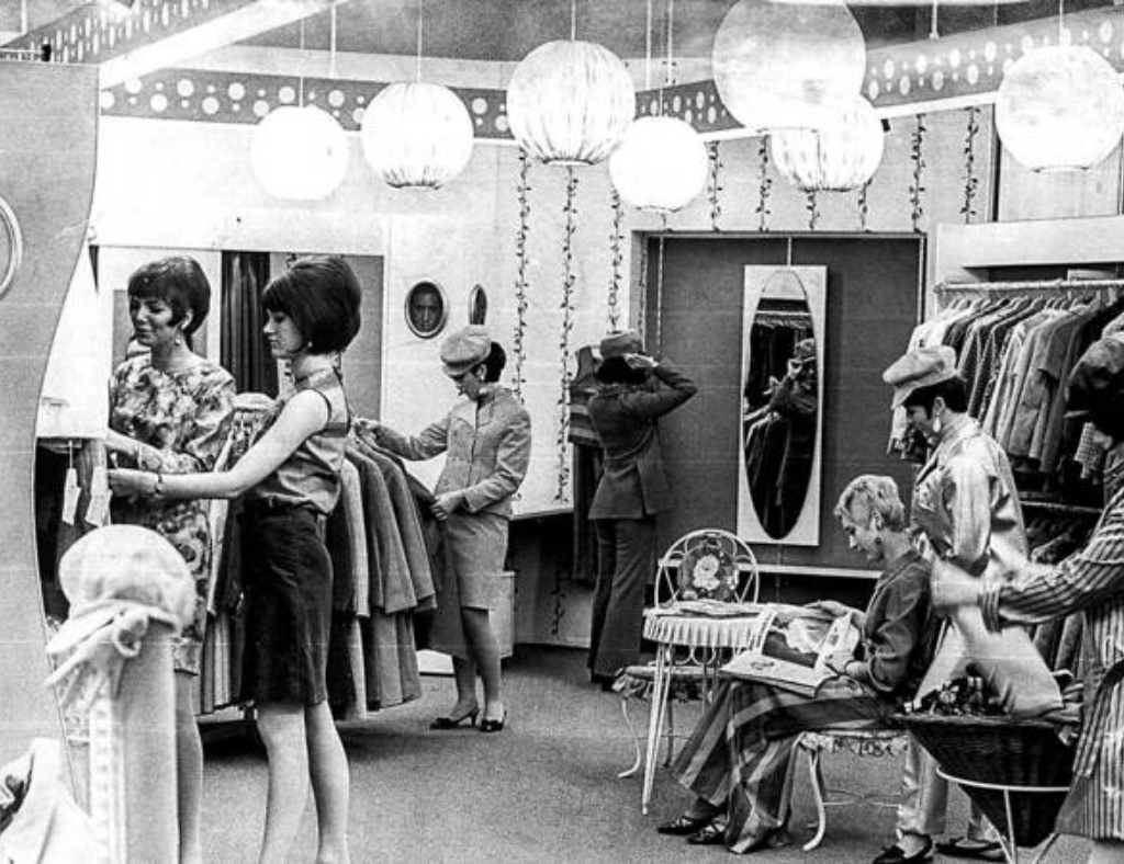 Schwarz-Weiß-Foto eines Retro-Modegeschäfts. Mehrere Frauen stöbern in Kleiderständern, während andere sitzen. Zur Dekoration gehören hängende Laternen und Lichterketten. Ein Spiegel an der Wand reflektiert einen Teil der Szene.