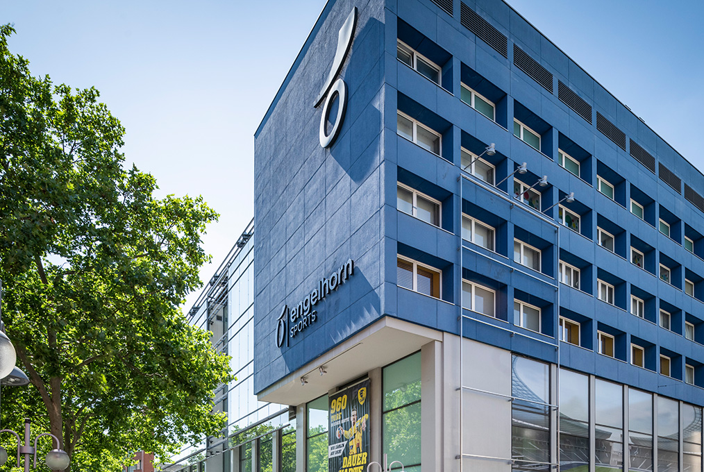 Ein blaues Gebäude mit dem Logo von Engelhorn Sports. Das Gebäude verfügt über mehrere Fenster und ist teilweise von Bäumen unter klarem Himmel umgeben.
