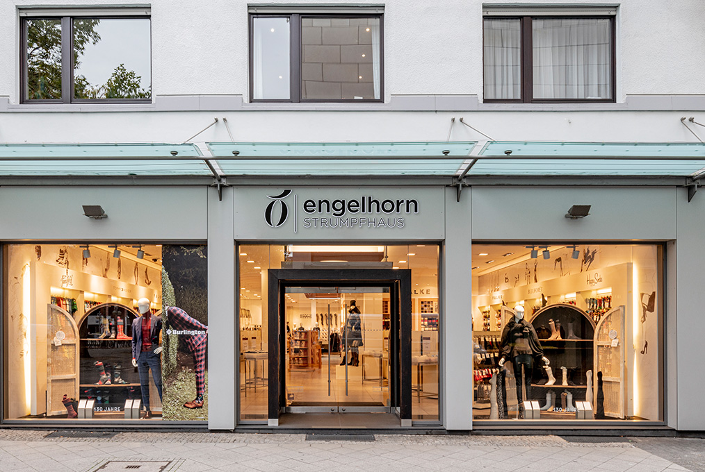 Schaufensterfront des Engelhorn Strumpfhauses mit großen Glasfenstern, in denen Schaufensterpuppen in Sportbekleidung und Accessoires ausgestellt sind. Der Eingang befindet sich in der Mitte, das Gebäude ist grau mit einer blaugrünen Markise.