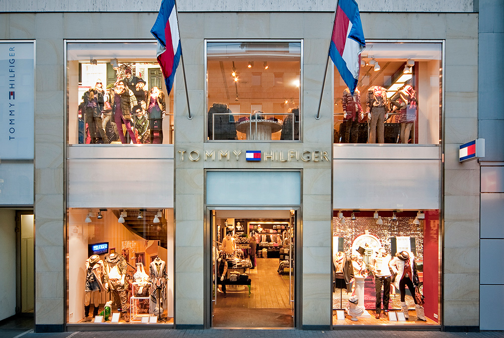 Zweistöckige Fassade des Tommy Hilfiger Stores mit großen Fenstern, in denen Schaufensterpuppen in verschiedenen Outfits ausgestellt sind. Darüber hängen zwei Flaggen, und das Markenlogo ist mittig zwischen den Etagen platziert. Im Inneren sind Regale und weitere Kleidungsstücke zu sehen.