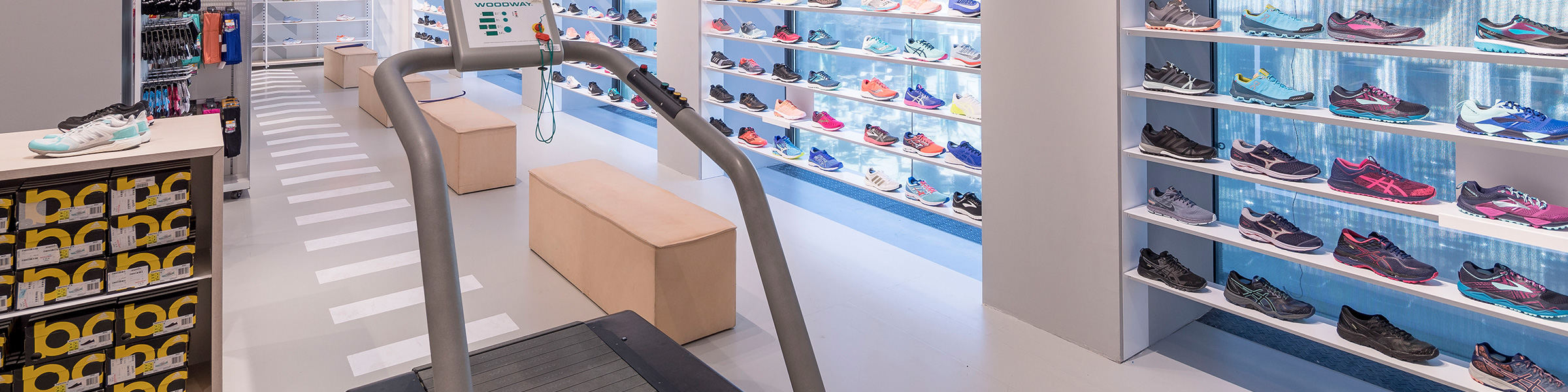 Ein Ladeninneres mit mehreren Reihen Sportschuhen, die auf weißen Regalen an den Wänden präsentiert werden. Beige Bänke stehen in der Mitte, im Vordergrund ein Laufband. Helles Licht hebt die Schuhkollektion hervor.