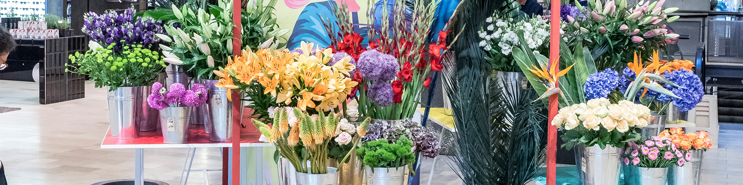 Eine farbenfrohe Präsentation verschiedener Blumen in Metalleimern, darunter orangefarbene Lilien, rote Gladiolen, violette Nelken, weiße Rosen und Paradiesvögel, vor einem farbenfrohen Hintergrund in einem Laden.