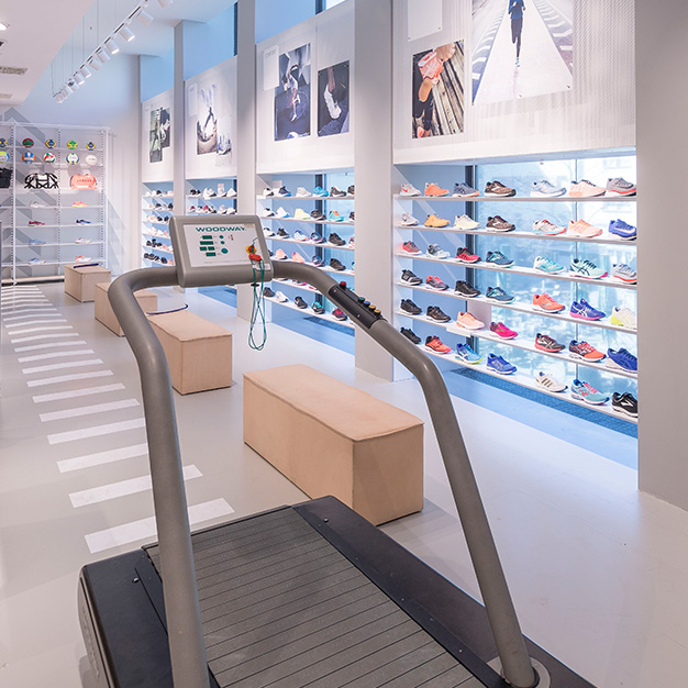 Innenansicht eines modernen Schuhgeschäfts mit einer Auswahl an Turnschuhen in den Regalen. Im Vordergrund steht ein Laufband mit Digitalanzeige. Das Geschäft besticht durch elegante Designelemente und große Fotos über den Regalen.