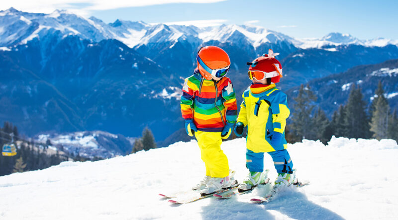 Zwei Kinder, die auf Skiern in Schneekleidung auf einem Berg stehen.