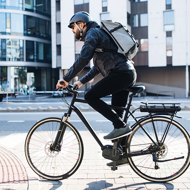 Eine Person fährt mit dem Fahrrad durch die Stadt. Sie trägt Helm, Sonnenbrille und Rucksack. Sie radelt an einem sonnigen Tag an modernen Gebäuden entlang.