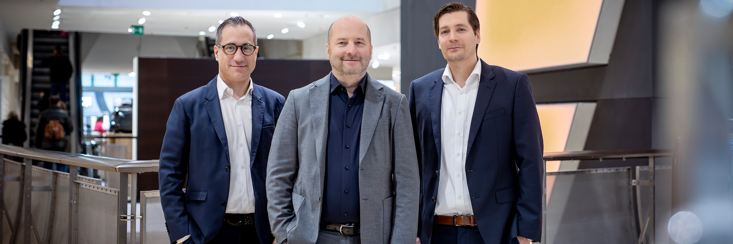 Drei Männer in Businesskleidung stehen in einem modernen Innenraum. Der Mann in der Mitte trägt einen hellgrauen Anzug, die beiden anderen dunkle Anzüge. Sie lächeln, mit warmem Licht und architektonischen Elementen im Hintergrund.