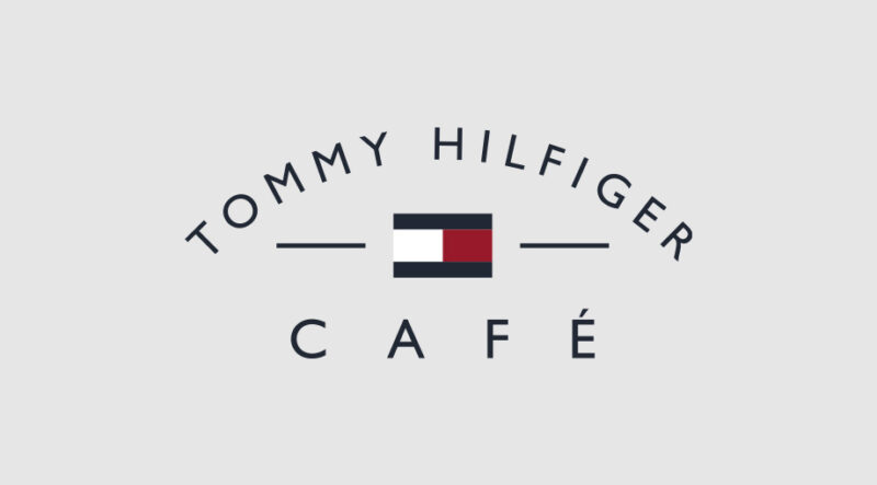 Logo für „Tommy Hilfiger Café“ mit dem Markenemblem in Rot, Weiß und Blau zwischen dem Text. Die Worte „Tommy Hilfiger“ sind oben gebogen, „Café“ steht darunter auf hellgrauem Hintergrund.
