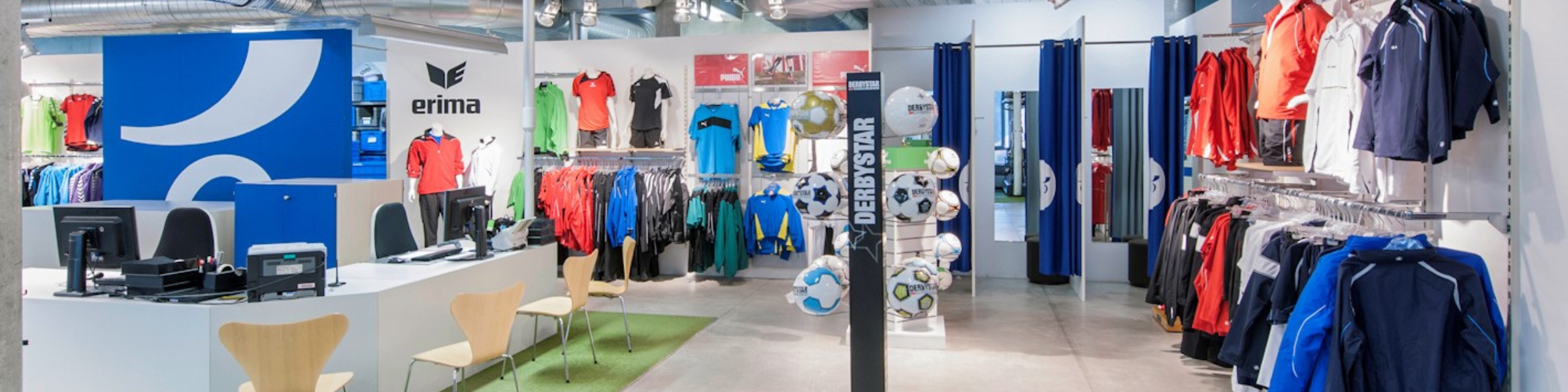 Ein Sportgeschäft mit Regalen voller bunter Sportbekleidung an den Wänden. Links befindet sich eine weiße Theke mit Computern und Holzstühlen. In der Mitte sind Fußbälle und Sportausrüstung ausgestellt. Helles Licht erhellt den Raum.
