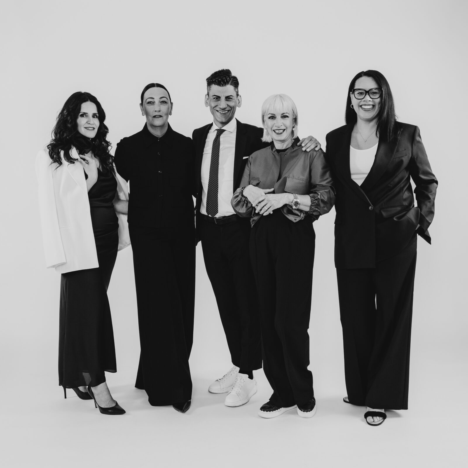 Eine Gruppe von fünf Personen, vier Frauen und ein Mann, steht in einem Studio zusammen. Alle sind formell gekleidet, die Frauen tragen Kleider und Blazer, der Mann einen Anzug. Sie lächeln und stehen entspannt vor einem schlichten Hintergrund.