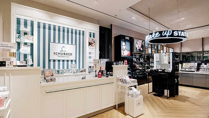 Ein gut beleuchteter Kosmetikladen mit einem weißen Tresen links mit der Aufschrift „SCHUBACK Parfümerie“. Rechts sind Regale voller Make-up-Produkte zu sehen, unter einem leuchtenden Schild „Make Up Station“. Der Boden ist aus hellem Holz.