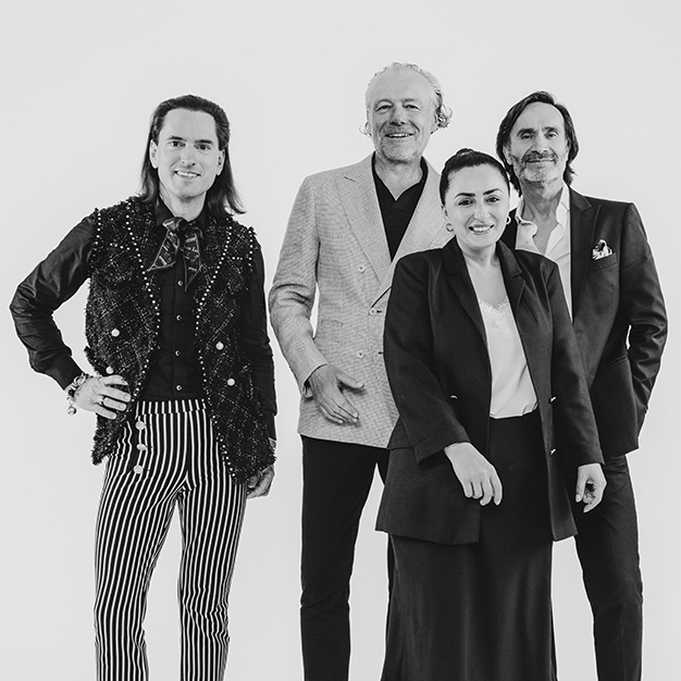 Vier Personen stehen vor einem weißen Hintergrund zusammen. Eine Person trägt eine Weste und gestreifte Hosen, zwei tragen Anzüge und eine einen Blazer mit langem Rock. Sie scheinen alle zu lächeln. Das Bild ist schwarz-weiß.