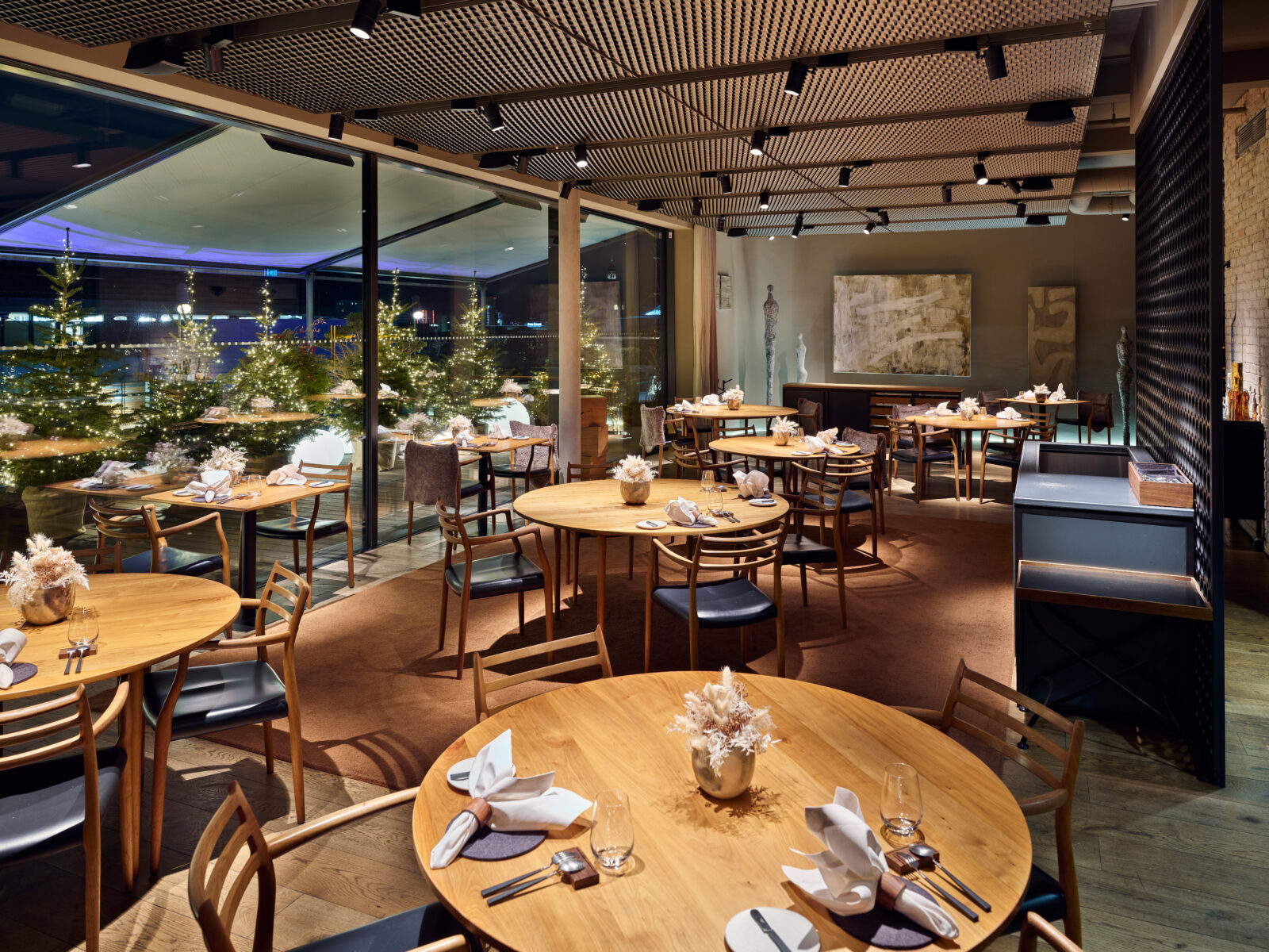 Ein moderner Restaurant-Essbereich mit Holztischen, weißen Servietten und Gläsern. Große Fenster bieten einen Blick auf beleuchtete Bäume bei Nacht. Die Einrichtung ist elegant, mit neutralen Tönen und sanfter Beleuchtung.