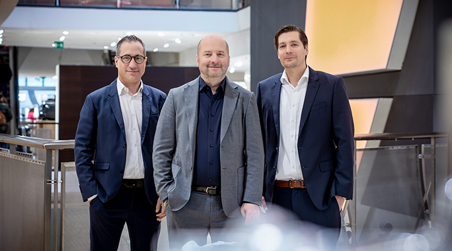 Drei Männer in Businesskleidung stehen in einer modernen Büroumgebung zusammen. Zwei tragen dunkle Anzüge, einer einen grauen. Sie wirken selbstbewusst und professionell und stehen neben einem Geländer mit sanft beleuchtetem Hintergrund.