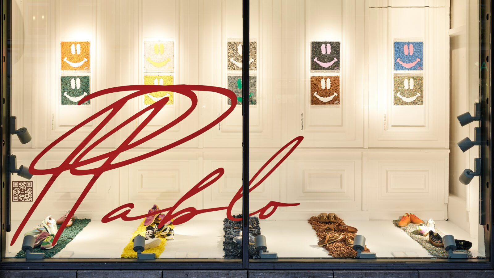 Schaufenster mit farbenfrohen Smiley-Kunstwerken und gewebten Gegenständen auf dem Boden. Der große rote Schriftzug „Pablo“ prangt quer über dem Fenster. Der Hintergrund ist weiß, mit gerahmten Kunstwerken in einem Raster angeordnet.