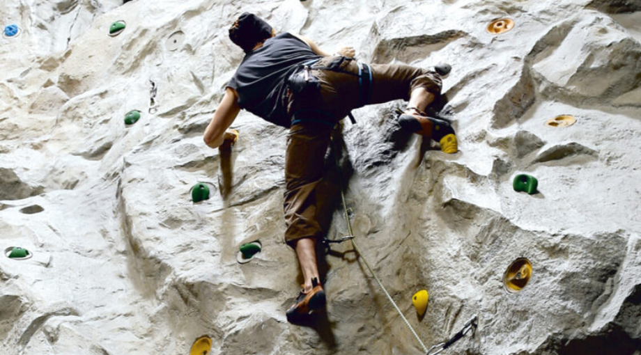 Eine Person mit einem Klettergurt klettert eine Indoor-Kletterwand hinauf und nutzt dabei bunte Hand- und Trittgriffe, die an der strukturierten Oberfläche befestigt sind.