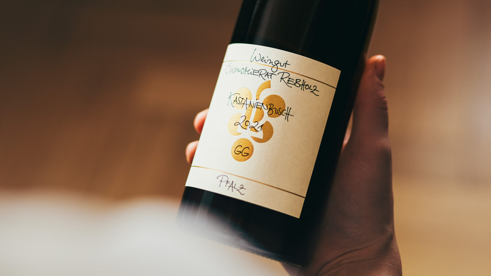 Eine Hand hält eine dunkle Weinflasche mit einem cremefarbenen Etikett, auf dem handschriftlicher Text steht, darunter „Weingut“, „2021“, „GG“ und „Pfalz“. Der Hintergrund ist unscharf.