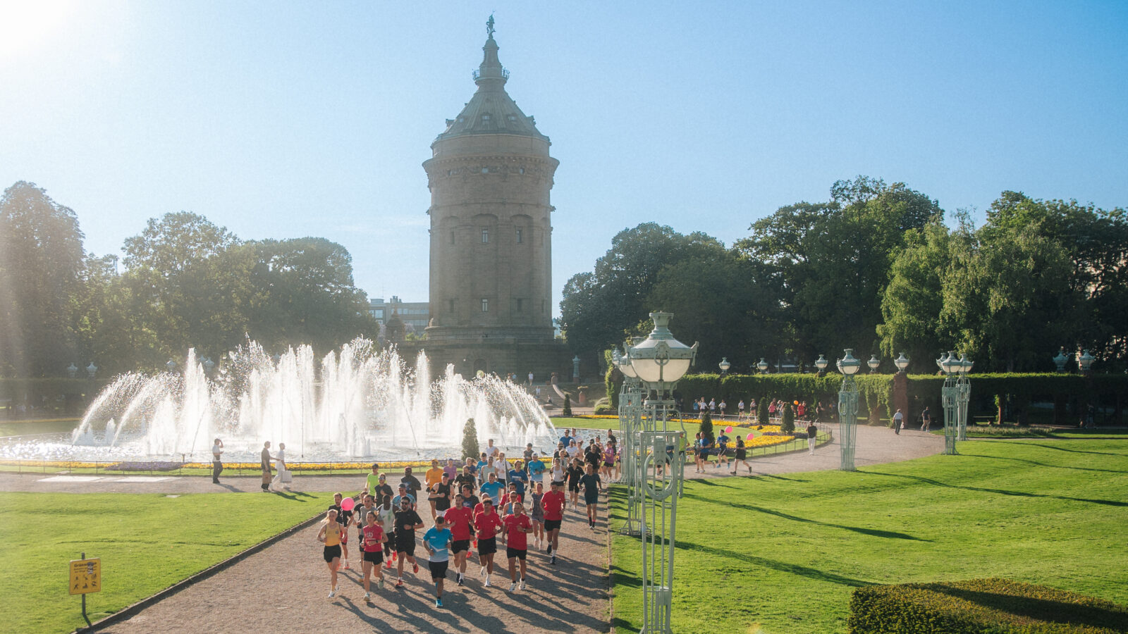 Eine Gruppe Läufer läuft an einem großen Brunnen in einem sonnigen Park vorbei. Im Hintergrund ein historischer Turm und gepflegte Rasenflächen. Der Himmel ist klar und blau, Bäume säumen den Weg.
