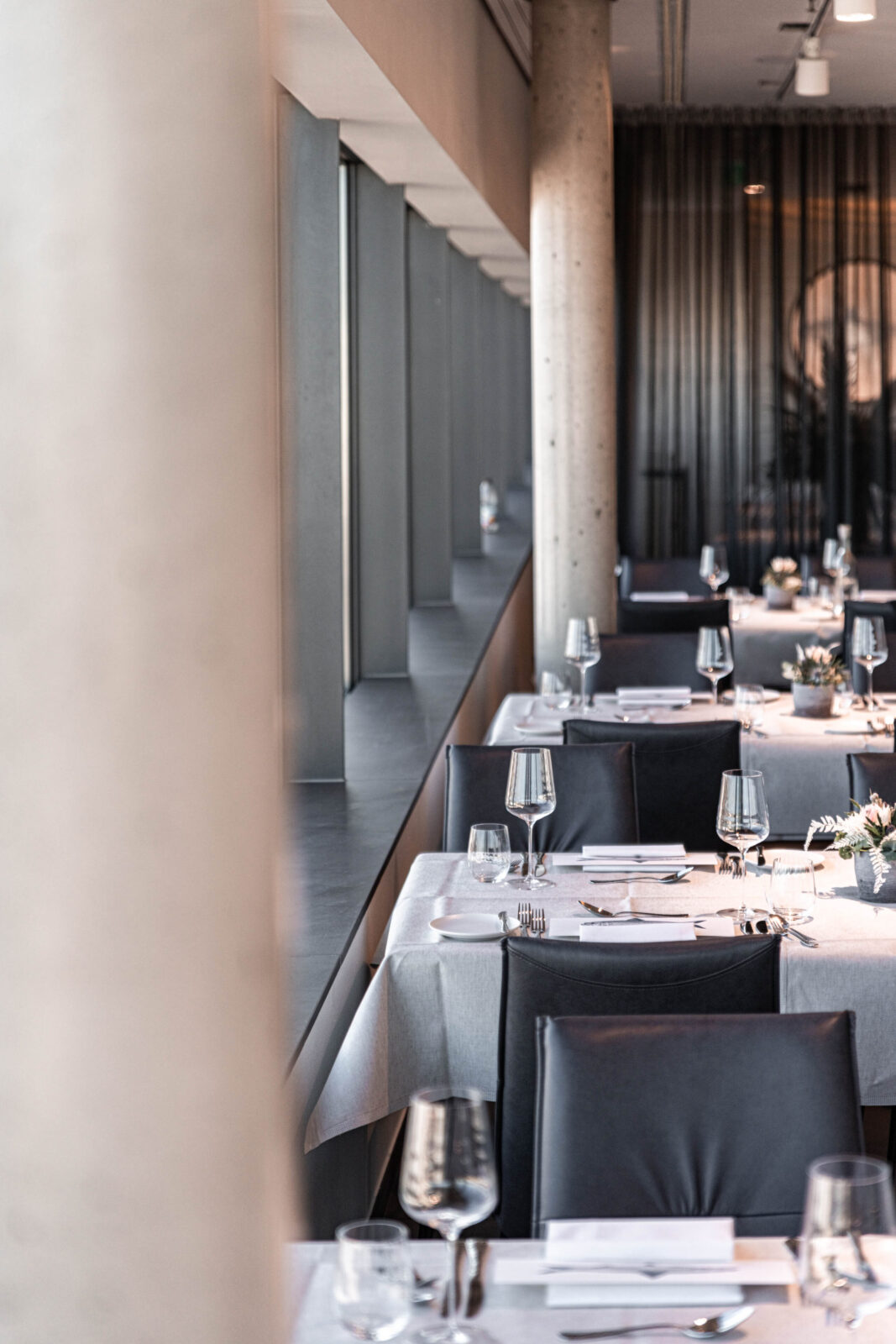 Elegantes, modernes Restaurant-Interieur mit schwarzen Stühlen, weißen Tischdecken und hübsch gedeckten Tischen mit Weingläsern. Große Fenster an einer Seite lassen Tageslicht herein. Betonsäulen und transparente Vorhänge schaffen eine gehobene Atmosphäre.