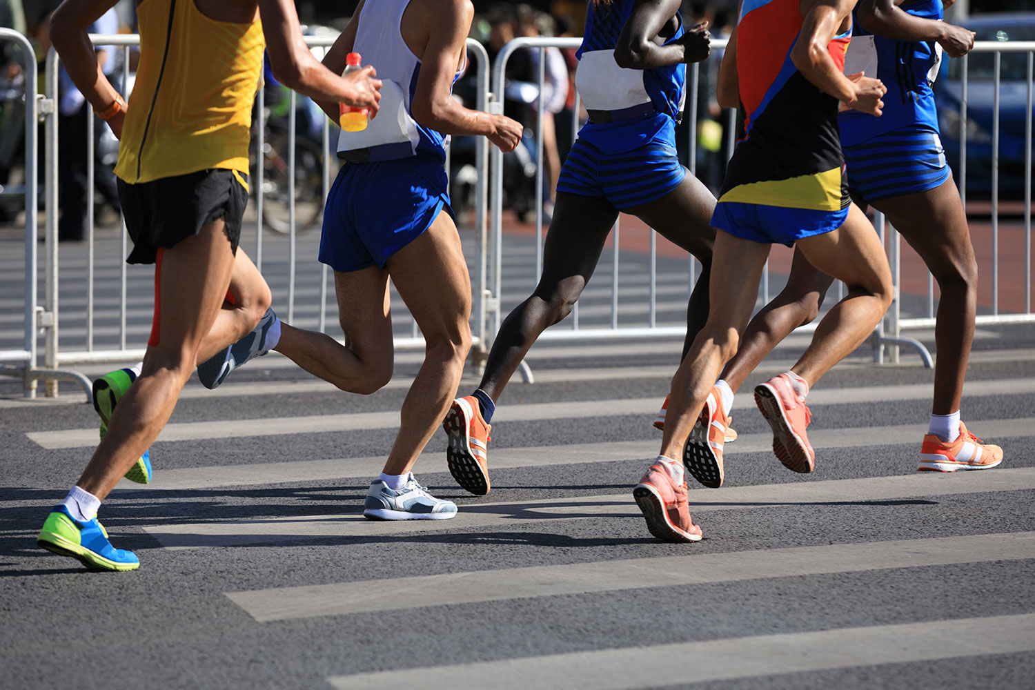 Eine Gruppe von Läufern in Sportkleidung, die an einem Straßenrennen teilnehmen, wurde mitten im Schritt aufgenommen, hinter ihnen sind Metallbarrieren und ein unscharfer Hintergrund zu sehen.