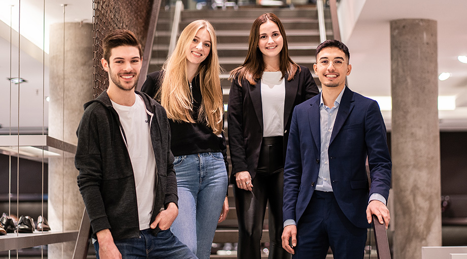 Vier junge Erwachsene, drei Frauen und ein Mann, stehen gemeinsam in einem Innenbereich vor einer Treppe. Sie lächeln, sind leger und geschäftsmäßig gekleidet und wirken in der modernen Umgebung selbstbewusst und freundlich.