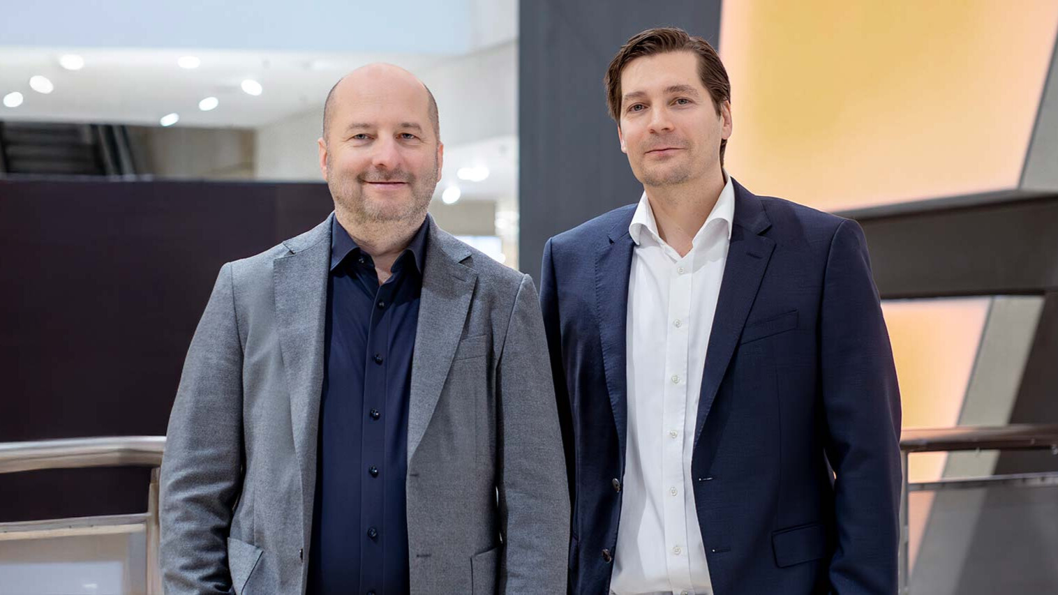Zwei Männer in Businesskleidung stehen in einem modernen Gebäude mit Glasgeländer und heller, sanfter Beleuchtung. Einer trägt einen grauen Blazer, der andere einen dunkelblauen Anzug. Beide blicken leicht lächelnd in die Kamera.