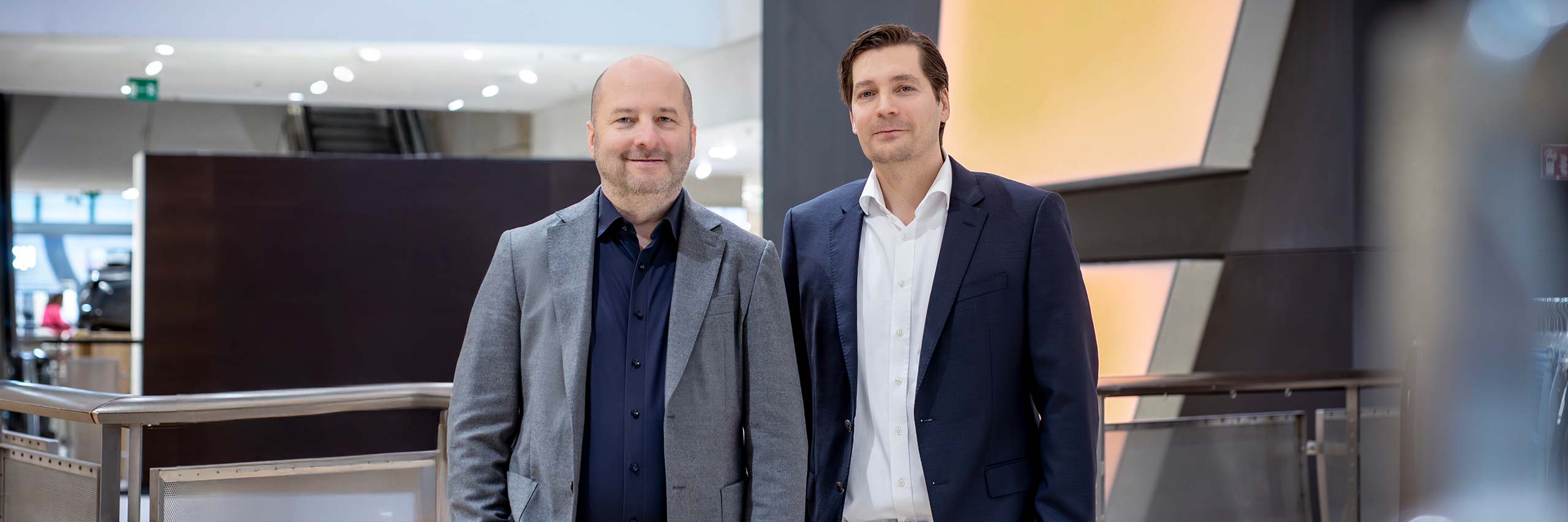 Zwei Männer in Businesskleidung stehen im Innenbereich und lächeln in die Kamera. Einer trägt einen grauen Blazer und ein dunkelblaues Hemd, der andere einen dunkelblauen Anzug und ein weißes Hemd. Im Hintergrund ist ein moderner, gut beleuchteter Innenraum zu sehen.