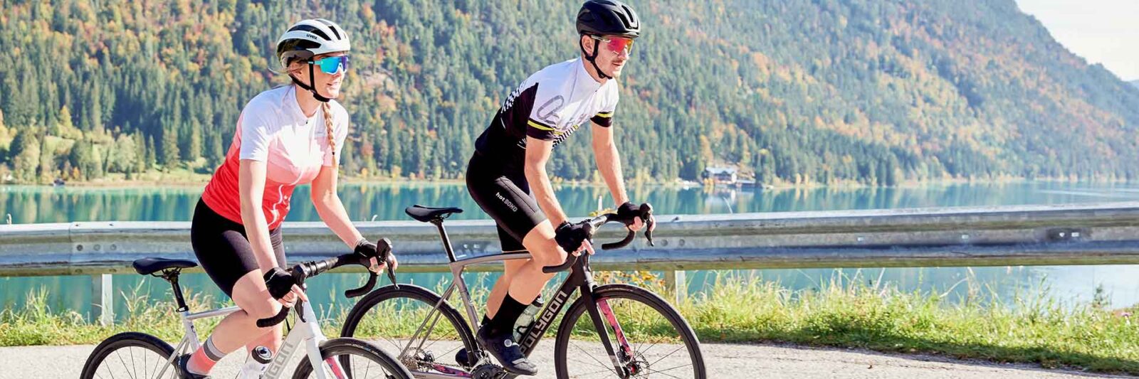 Zwei Radfahrer fahren mit Rennrädern eine Uferstraße entlang, im Hintergrund Berge und Bäume. Beide tragen Helme, Sonnenbrillen und Radbekleidung von Fahrrad engelhorn und genießen einen sonnigen Tag im Freien.
