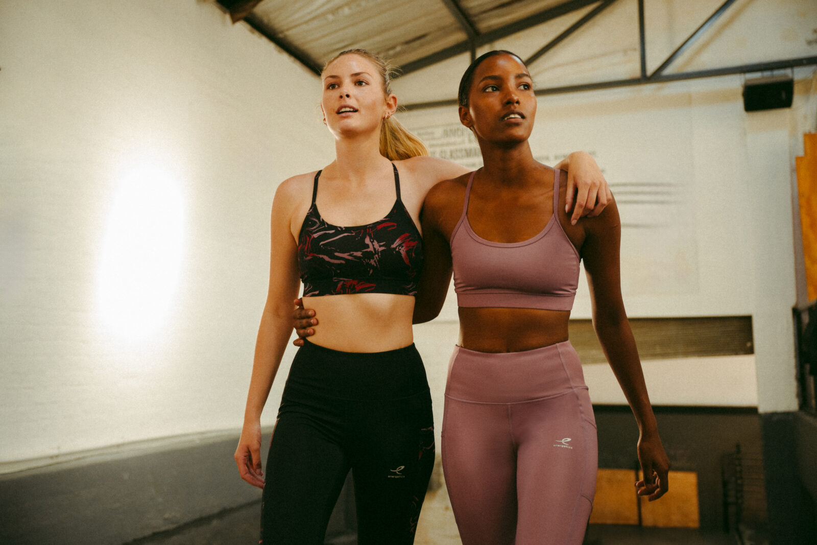 Zwei Frauen in Sportkleidung gehen Arm in Arm durch ein Fitnessstudio und blicken nach vorne. Die eine trägt einen schwarzen Sport-BH mit passenden Leggings, die andere einen lila Sport-BH und Leggings. Die Atmosphäre wirkt freundlich und unterstützend.