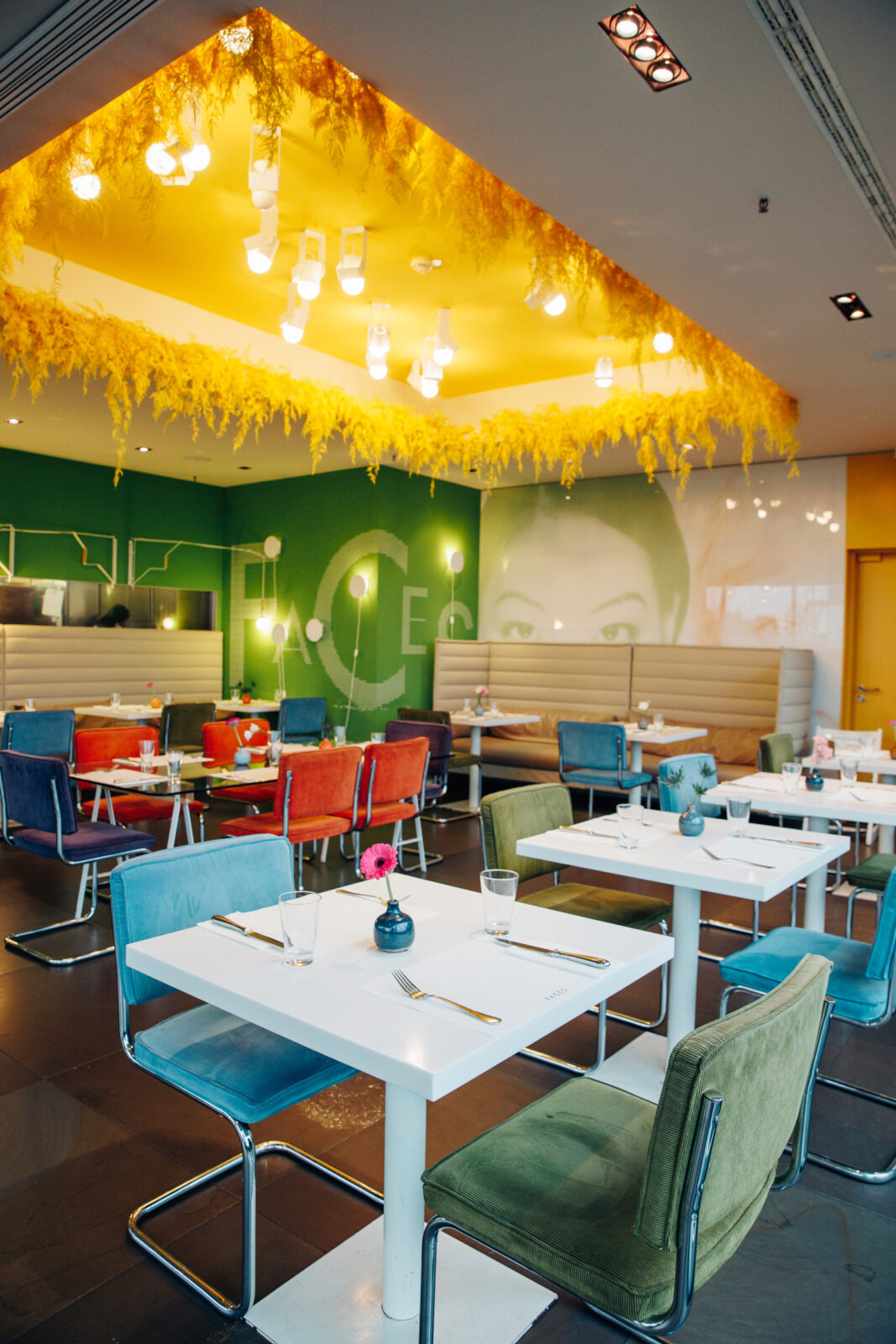 Ein modernes Restaurant mit farbenfrohen Stühlen, weißen Tischen und einer leuchtend grün-gelben Wand. Eine gelbe Decke mit Hängelampen und dekorativem Laub verleiht dem hellen, stilvollen Interieur Wärme.