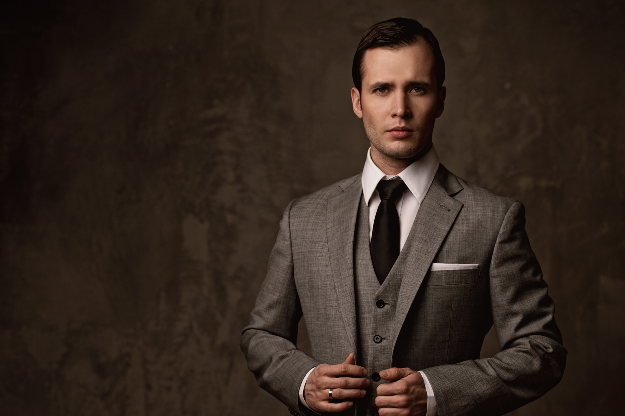 Junger Mann in beigem Anzug posiert ernst vor einem braunen Hintergrund.
